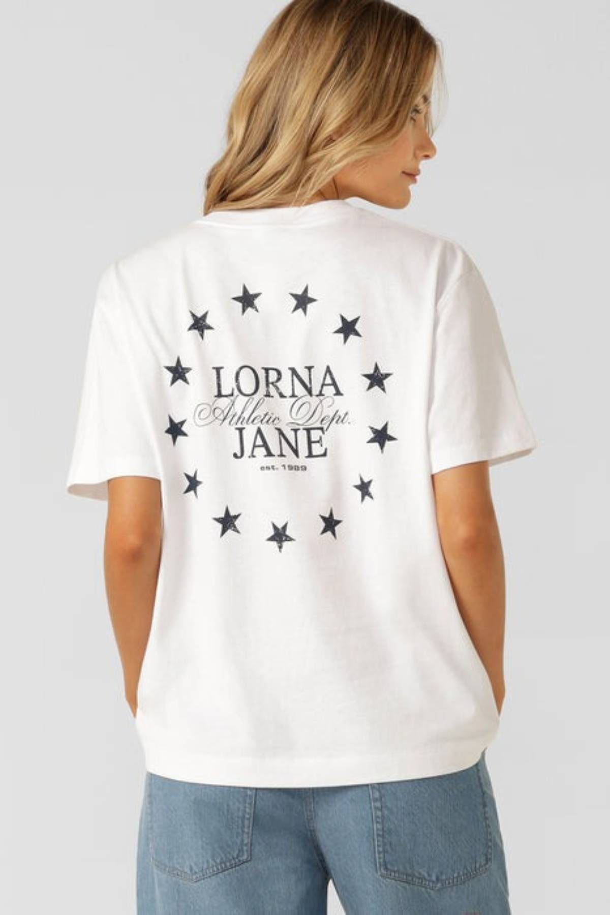 A woman faces away from the camera wearing the Lorna Jane Sprint Classic Fit T-Shirt—an oversized cotton jersey with "LORNA JANE Athletic Dept. est. 1989" and black stars on the back—paired with blue denim pants.