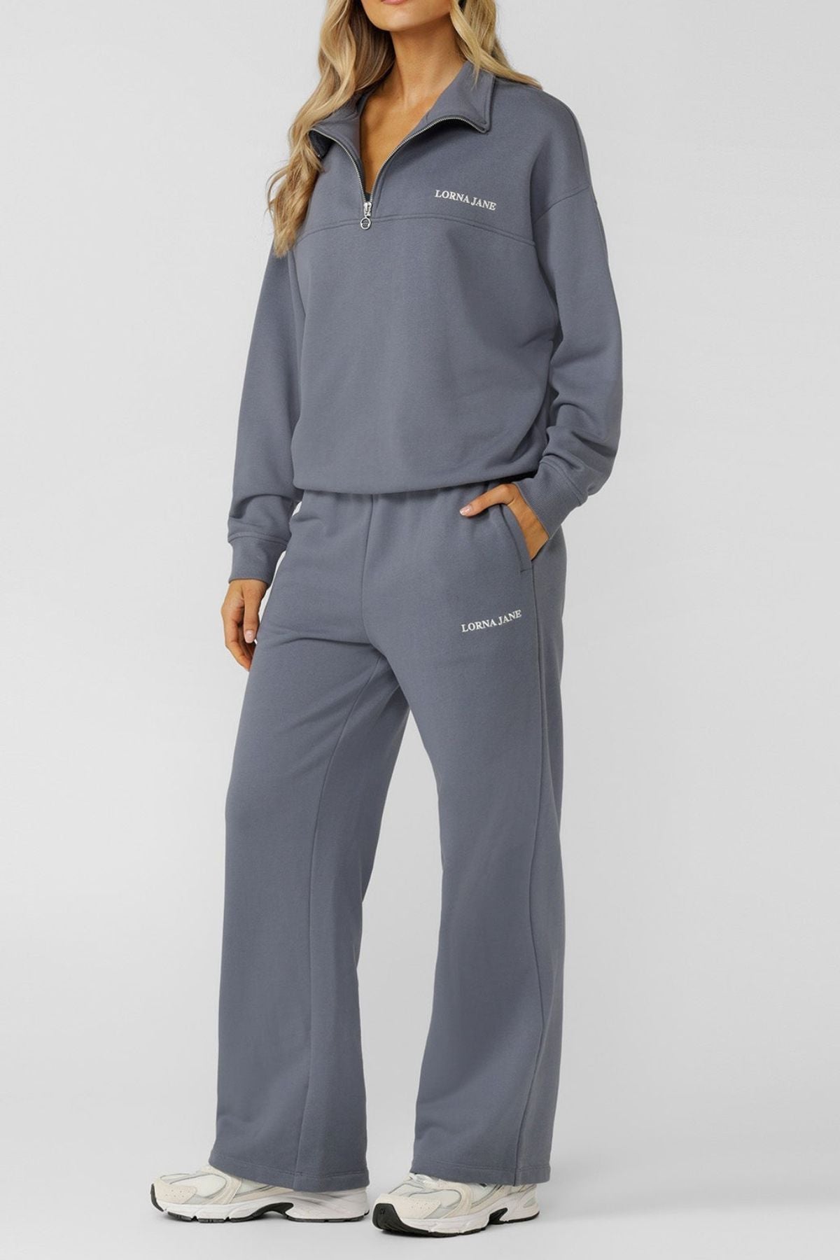 A woman with long, wavy blonde hair stands against a plain background, wearing the Lorna Jane Streamline Quarter Zip Sweat and matching gray wide-leg pants with "Lorna Jane" text, paired with white sneakers.
