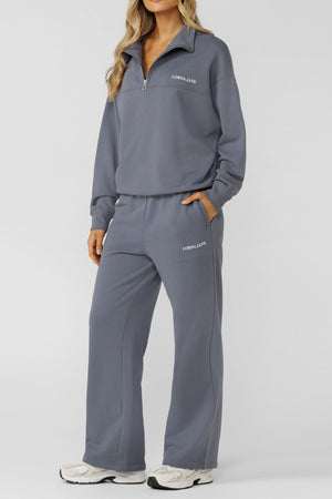 A woman with long, wavy blonde hair stands against a plain background, wearing the Lorna Jane Streamline Quarter Zip Sweat and matching gray wide-leg pants with "Lorna Jane" text, paired with white sneakers.
