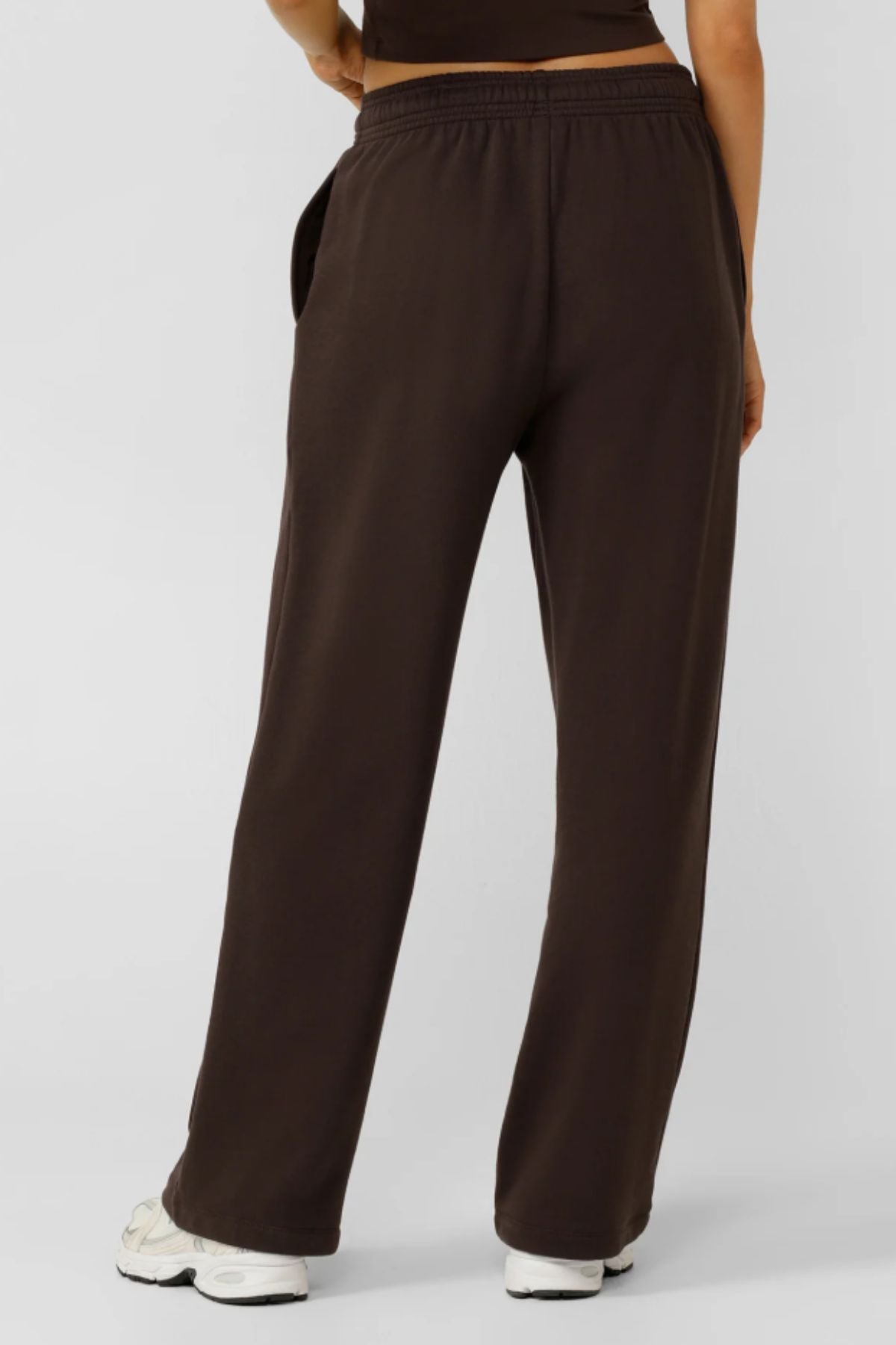 A person stands facing away, wearing Lorna Jane Streamline Sweatpants in dark brown with white sneakers and a partially visible dark shirt against a plain, light background.