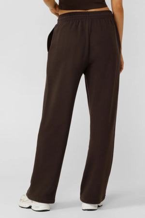 A person stands facing away, wearing Lorna Jane Streamline Sweatpants in dark brown with white sneakers and a partially visible dark shirt against a plain, light background.