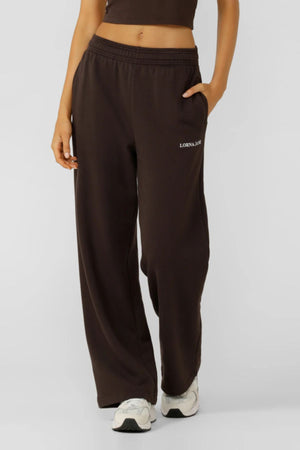 A person in dark brown Lorna Jane Streamline Sweatpants with "LORNA JANE" on the left thigh, a matching cropped top, and white sneakers—stylish, comfortable women’s lounge pants for effortless everyday wear.