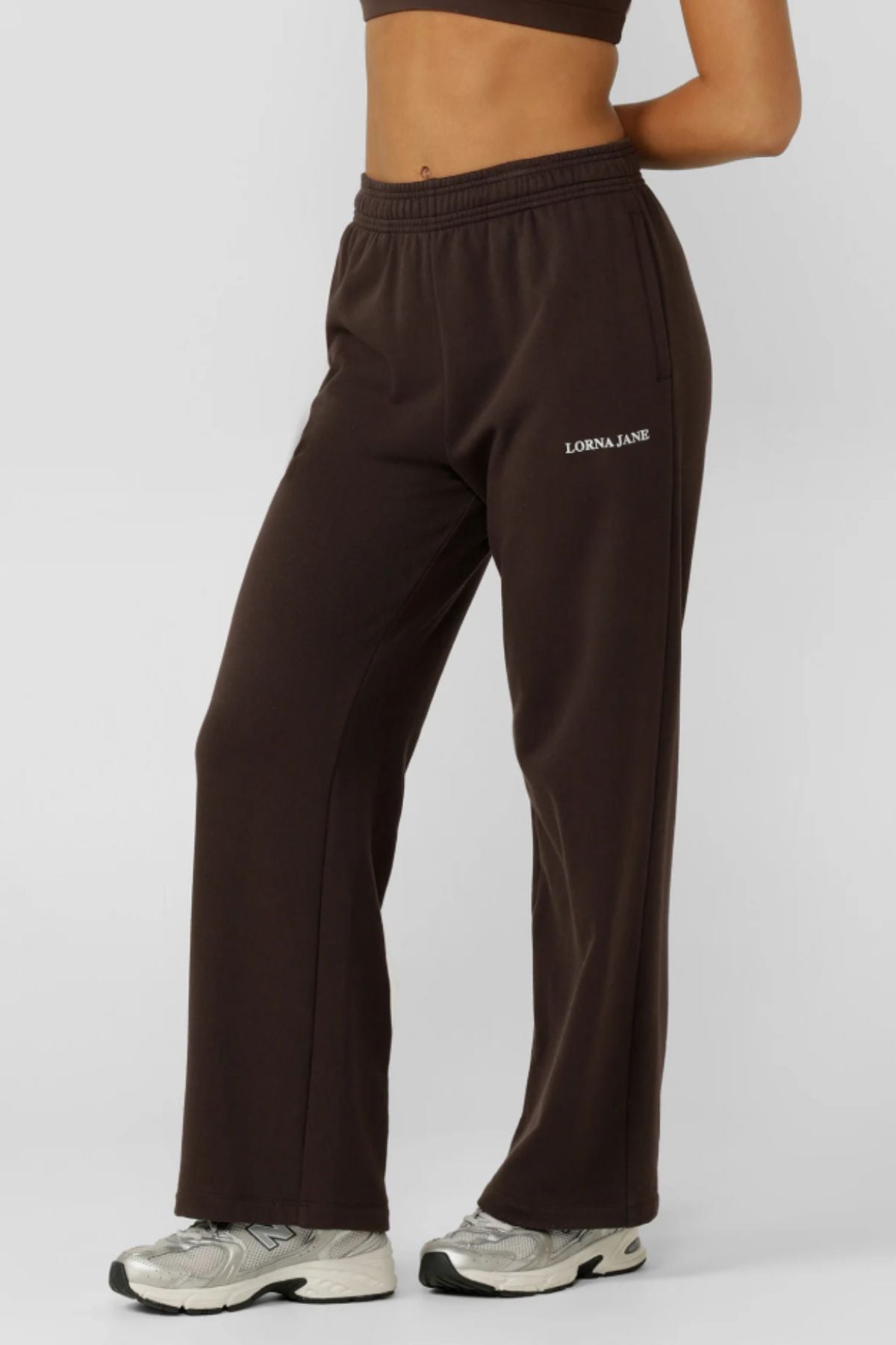 A person wearing dark brown Lorna Jane Streamline Sweatpants and silver athletic shoes stands against a plain light background, with only the lower torso and legs visible to highlight the sweatpants’ relaxed fit style.