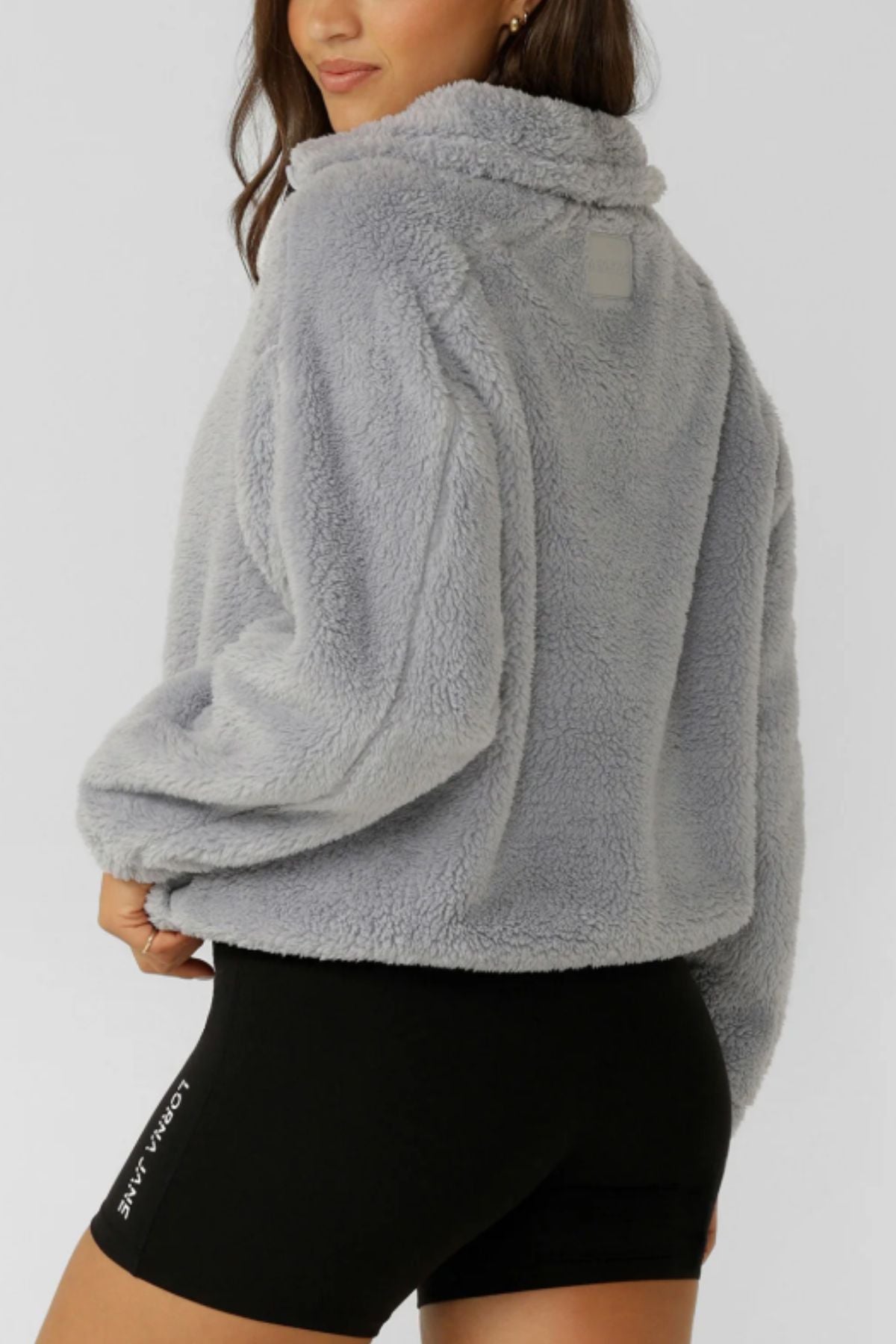 A person wearing the Lorna Jane Everyday Teddy Quarter Zip Sweat in light gray and black fitted shorts with "Lorna Jane" on the leg, posing with one hand on their hip against a plain background.
