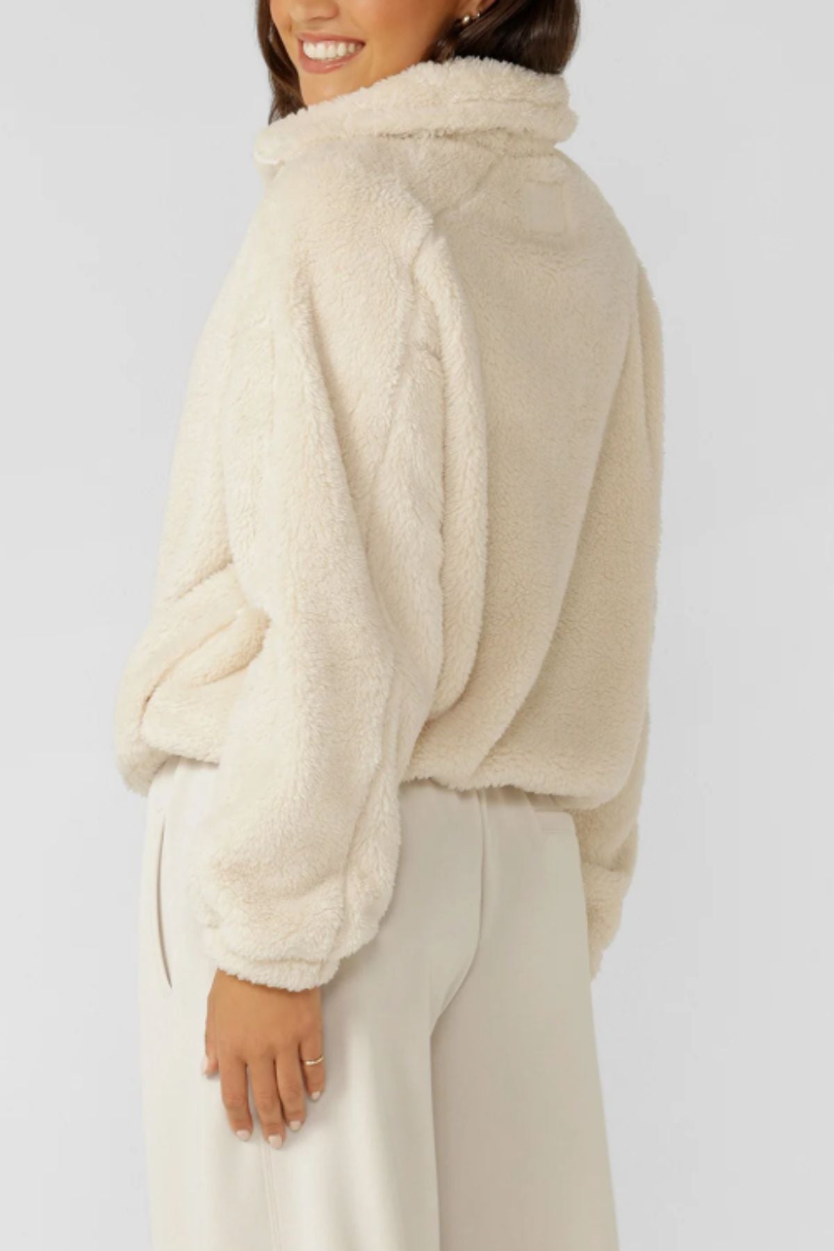 A person wearing a cream Lorna Jane Everyday Teddy Quarter Zip Sweat with an oversized fit and light pants stands sideways, smiling with one hand in their pocket against a plain background.