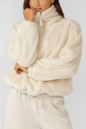 A person wearing a Lorna Jane Everyday Teddy Quarter Zip Sweat, a cosy oversized women's sweatshirt with a high collar, and matching cream drawstring pants stands with one hand touching their face.