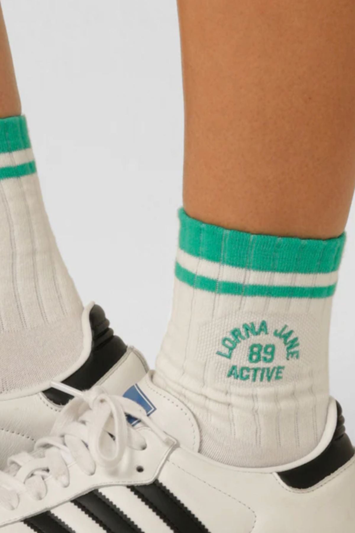 Close-up of a person wearing Lorna Jane Throwback Crew Socks, white cotton with green stripes and "Lorna Jane Active 89" text, paired with white and black Adidas sneakers.