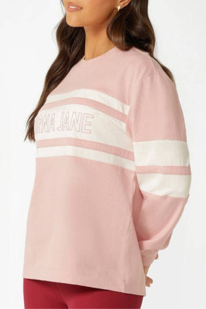 A woman wears the Lorna Jane Trackside Long Sleeve T-Shirt in light pink with white stripes and "LORNA JANE" text, styled with burgundy leggings. She stands sideways, hands behind her back.