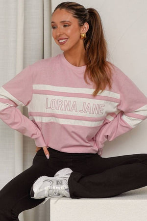 A woman with long brown hair in a ponytail wears the Lorna Jane Trackside Long Sleeve T-Shirt in pink and white, black pants, and white sneakers. She is smiling and sitting cross-legged on a white surface.