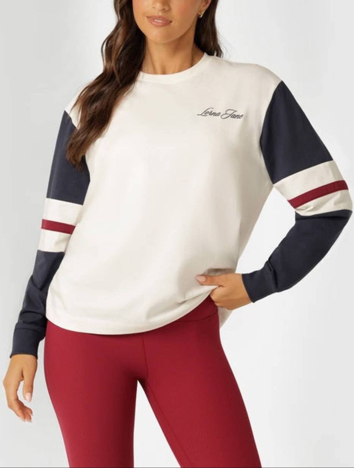 A woman stands with one hand on her hip, looking slightly to the side, wearing a white Lorna Jane Varsity Long Sleeve T-Shirt with navy and burgundy striped sleeves and red leggings.