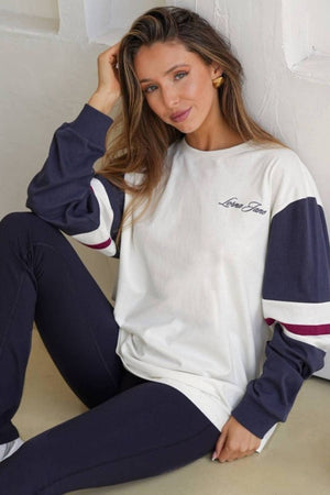 A woman with long, light brown hair sits on the floor wearing a Lorna Jane Varsity Long Sleeve T-Shirt and navy leggings. Made from breathable cotton, her white and navy top features โLove Geneโ as she smiles at the camera.
