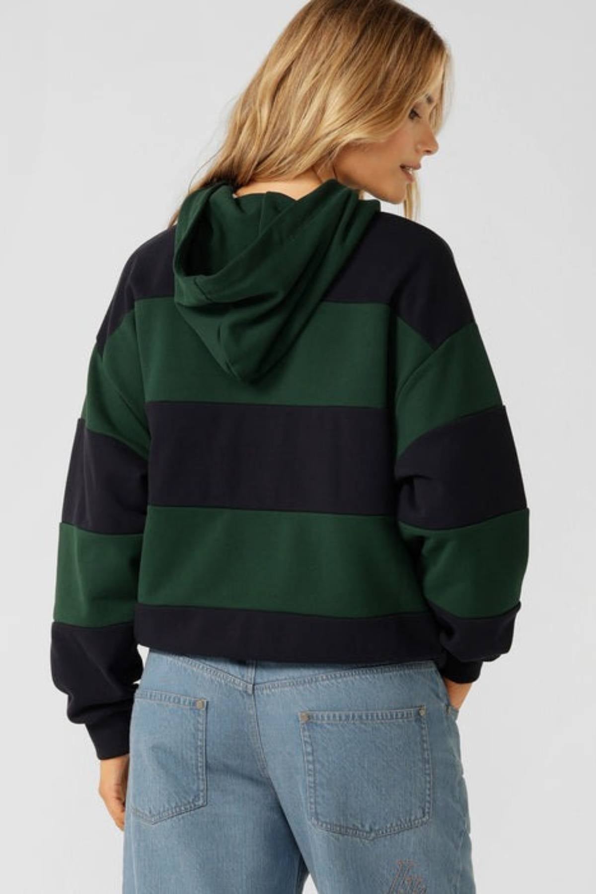 A person with long blonde hair is seen from the back in a Lorna Jane Heritage Rugby Polo Hoodie with dark green and black stripes, paired with light blue jeans, slightly turned to the side against a plain background.
