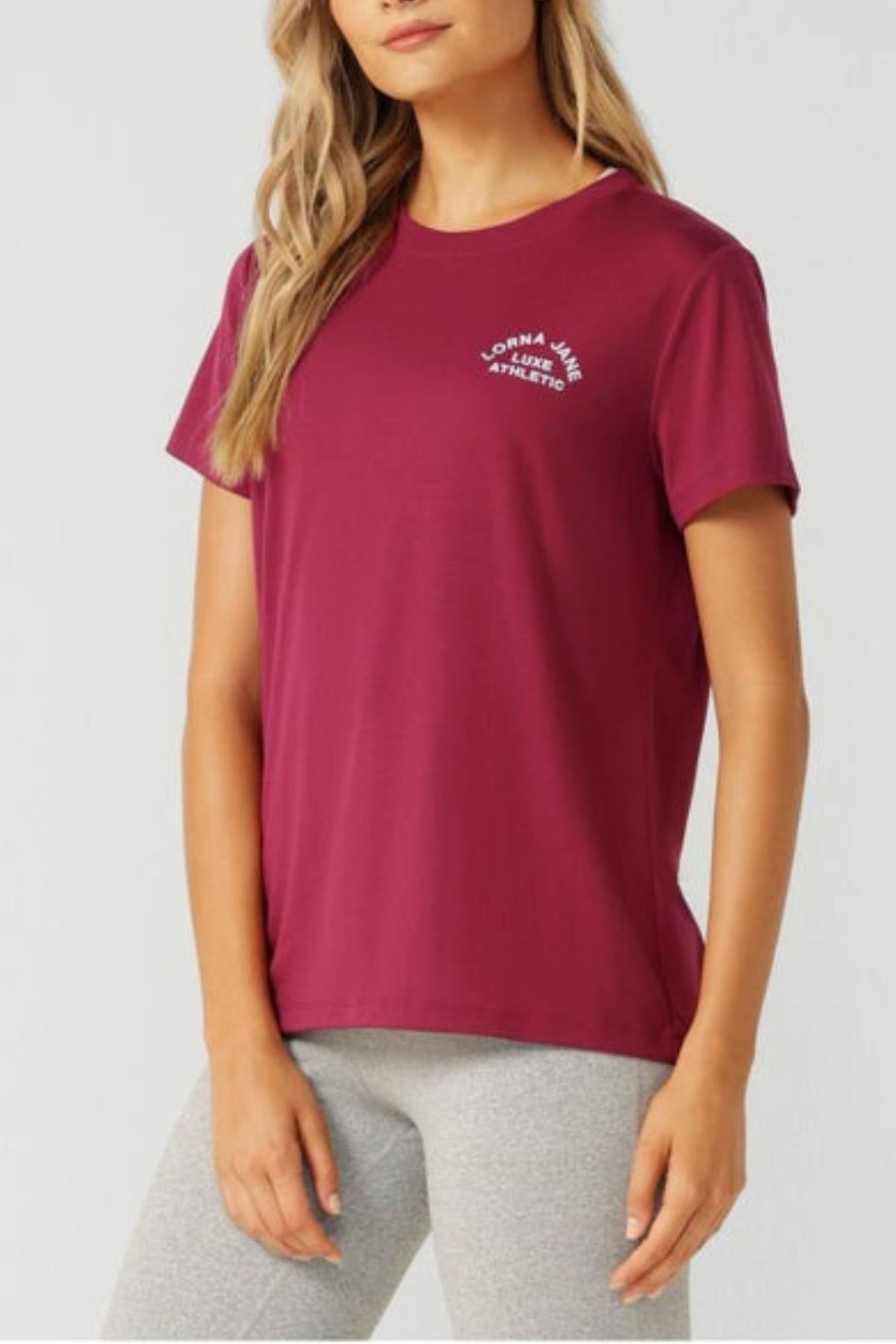 A woman wears the Lorna Jane Lotus T-Shirt with white text on the left chest and light gray leggings, standing against a plain light background—ideal for women’s activewear and an active lifestyle.
