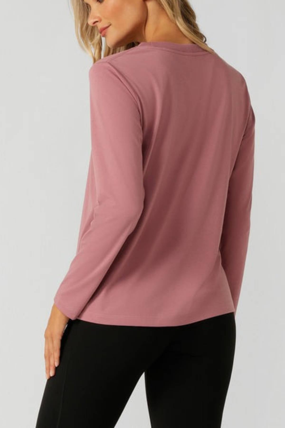 A blonde woman stands with her back to the camera, head turned slightly, wearing the Lorna Jane Lotus Long Sleeve T-Shirt in mauve and black pants, highlighting the activewear’s breathable fabric.