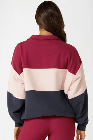 A woman with long brown hair faces away, wearing the Lorna Jane Sidelines Panelled Quarter Zip Sweat in maroon, light pink, and dark blue stripes, paired with maroon pants against a plain light background.