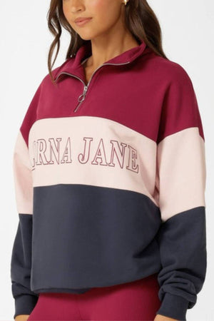 A woman wears the Lorna Jane Sidelines Panelled Quarter Zip Sweat, an oversized French terry cotton sweatshirt in maroon, light pink, and navy blue with "CARNA JANE" on the chest.