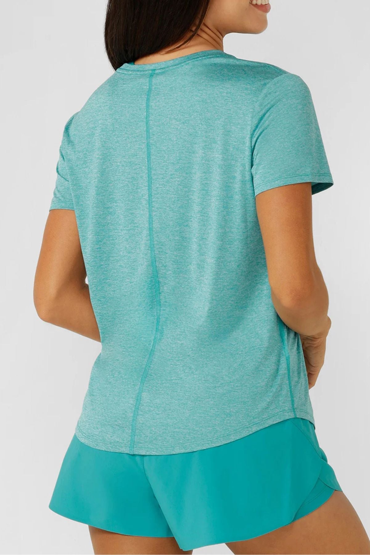 A woman, seen from the back, wears a light teal Lorna Jane Amy Dynamic Active T-Shirt and teal shorts. With one hand on her hip, she smiles slightly against a plain background. The workout top features breathable fabric for comfort.