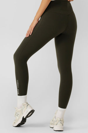 A person stands facing away, wearing dark green Lorna Jane Lotus No Chafe Phone Pocket Ankle Biter Leggings with "LORNA JANE" printed near the ankle, paired with white socks and sneakers—showcasing stylish activewear.