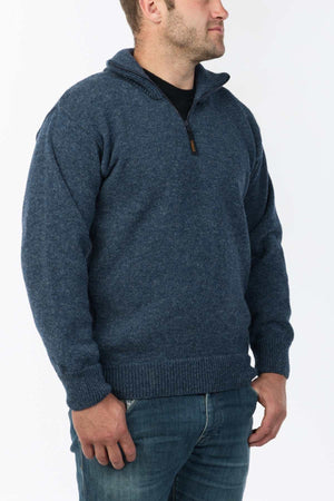 A man wearing the MKM North Wester Knitted Jumper in blue wool and blue jeans stands against a plain white background. He is facing slightly to the right with his hands relaxed by his sides. The jumper, made in New Zealand, features a ribbed collar, cuffs, and hem.