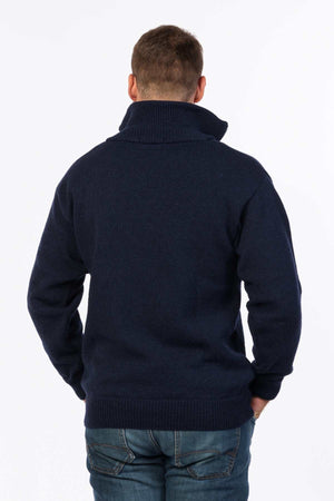 A person with short hair stands facing away from the camera, wearing a navy blue MKM North Wester Knitted Jumper made in New Zealand with the collar turned up and blue jeans. The person's right hand is in their pocket. The background is plain white.