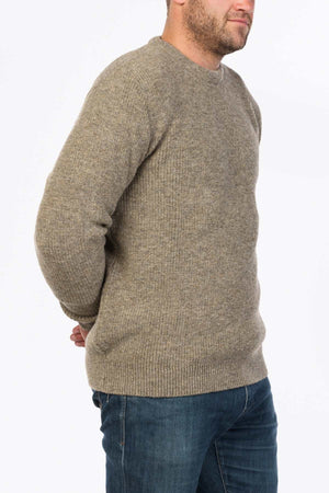 A man in the MKM Original Backyard Knit beige wool pullover and blue jeans stands against a plain white background, showcasing relaxed casual wear with his hands behind his back.