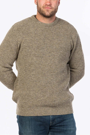 A person in the MKM Original Backyard Knit, a beige wool pullover, and blue jeans stands with their hands behind their back, set against a plain white background highlighting the simplicity of their casual look.