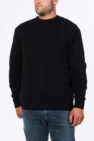 A person in a plain black MKM Original Backyard Knit pullover with a ribbed texture, long sleeves, and blue jeans stands against a white background. The person's arms are relaxed by their sides, showcasing the pullover's casual wear comfort.