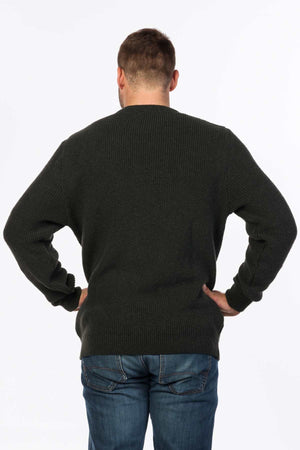 A person with short hair faces away, hands on hips, wearing an MKM Original Backyard Knit in dark green wool from New Zealand and blue jeans against a white background.