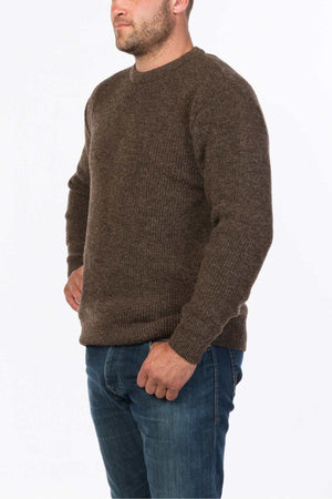 A man stands with his hand on his hip, clad in the MKM Original Backyard Knit, a brown ribbed pullover crafted from pure new wool, paired with blue jeans. He has a beard and a short haircut, all set against a plain white background.