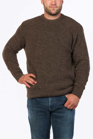 A person stands with one hand on their hip, wearing the MKM Original Backyard Knit pullover made of pure new wool in brown and blue jeans. The background is plain white.