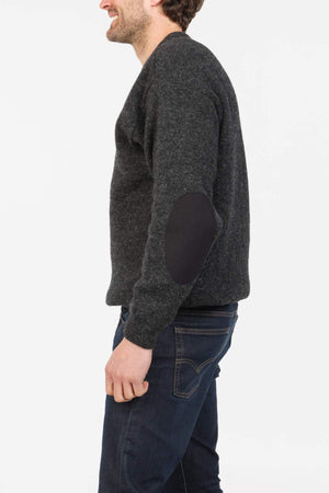 A man in profile wears the MKM Tasman Dual Layer Possum & Merino Crew sweater in dark gray with black elbow patches and dark blue jeans, standing against a plain white background.