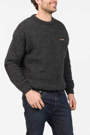 A man wears an MKM Tasman Dual Layer Possum & Merino Crew in dark grey, featuring double layer technology and a small chest logo, paired with dark blue jeans, as he stands smiling against a plain white background.
