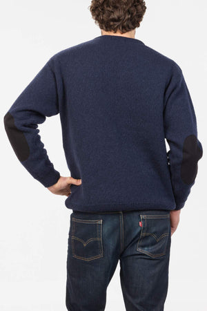 A person with curly hair, seen from the back, wears a navy blue MKM Tasman Dual Layer Possum & Merino Crew sweater with black elbow patches and dark blue jeans, standing with one hand on their hip against a white background.