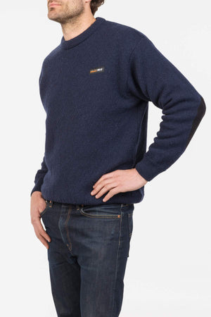 A man wearing the MKM Tasman Dual Layer Possum & Merino Crew in navy with black elbow patches and dark jeans stands with one hand on his hip against a white background, highlighting outdoor performance and style.