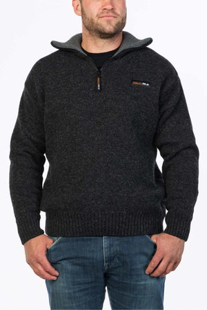 A man wearing an MKM Tasman Dual Layer Possum & Merino Knit, a dark gray zip-up sweater made of merino superfine wool with a high collar, stands with his left hand in the pocket of his blue jeans. The sweater features a small orange and white logo on the left chest. The background is plain white.