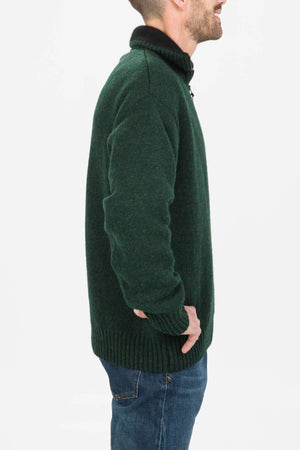 A man in a dark green MKM Tasman Dual Layer Possum & Merino Knit sweater and blue jeans stands in profile, facing right with his right hand in his pocket against a plain white background.