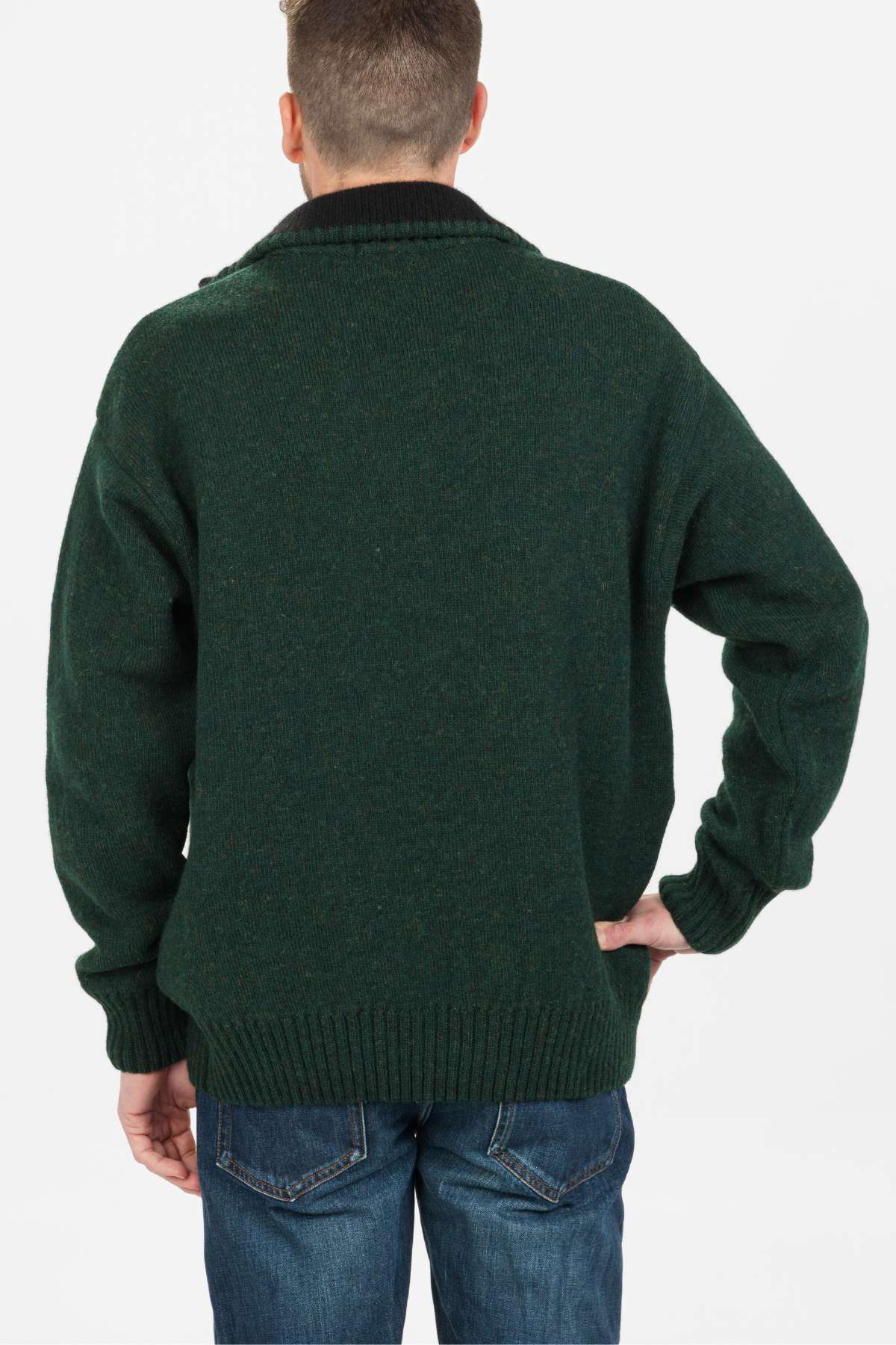 A person with short hair wearing a dark green MKM Tasman Dual Layer Possum & Merino Knit sweater and blue jeans stands facing away, one hand on their hip, against a plain white background.