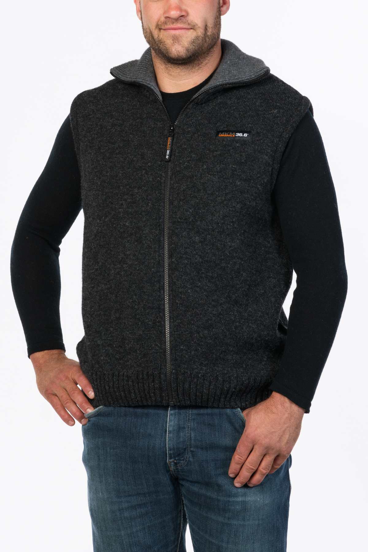 A person in a dark gray MKM Tasman Vest Dual Layer Possum & Merino over a black long-sleeve shirt and blue jeans stands with hands on hips against a plain white background.