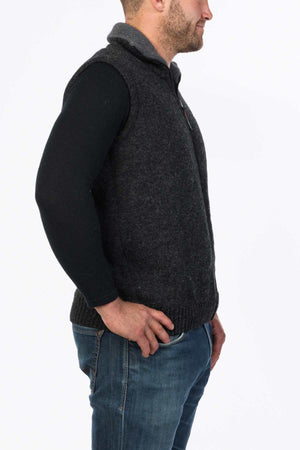 A man in profile, wearing an MKM Tasman Vest Dual Layer Possum & Merino over a black long-sleeve shirt and blue jeans, stands against a white background. His hands are in his pockets, and he has a short beard.