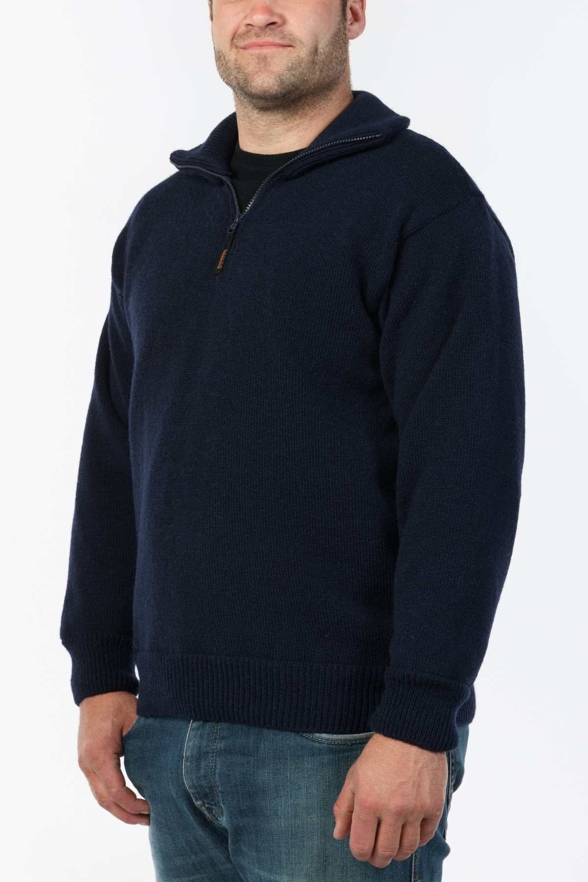 A person wearing the MKM Windbreaker Knit, a navy blue half-zip long-sleeve jumper crafted from 100% wool in New Zealand, stands against a light background. They smile slightly and pair it stylishly with blue jeans.