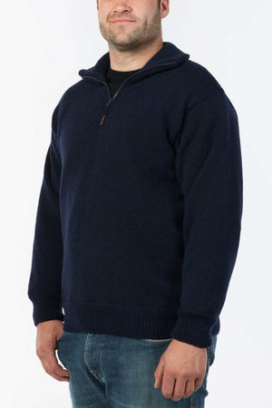A person wearing the MKM Windbreaker Knit, a navy blue half-zip long-sleeve jumper crafted from 100% wool in New Zealand, stands against a light background. They smile slightly and pair it stylishly with blue jeans.