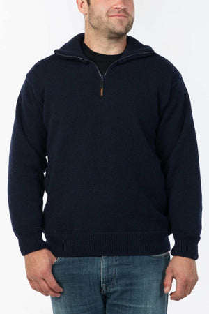 A man wearing the MKM Windbreaker Knit, a navy blue 100% wool jumper, over a black shirt and blue jeans stands against a plain white background, smiling slightly.