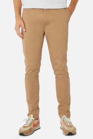 A person wearing Industrie The Cuba Chino Pant in a skinny fit beige stands against a plain background, paired with colorful sneakers featuring orange details and a light gray shirt, highlighting the casual elegance of their bottom half with hands resting in pockets.