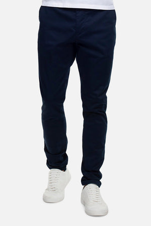 A person wearing dark blue Industrie The Cuba Chino Pants and white sneakers is shown from the waist down against a plain background. The slim-fit chinos paired with the simple, clean design of the white-laced sneakers exude a relaxed yet stylish vibe.