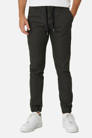 Someone models the Industrie The Drifter Chino Pant in slim-fit black with hands in pockets, paired with white sneakers and a white top, set against a plain white background, but only their lower half is visible.