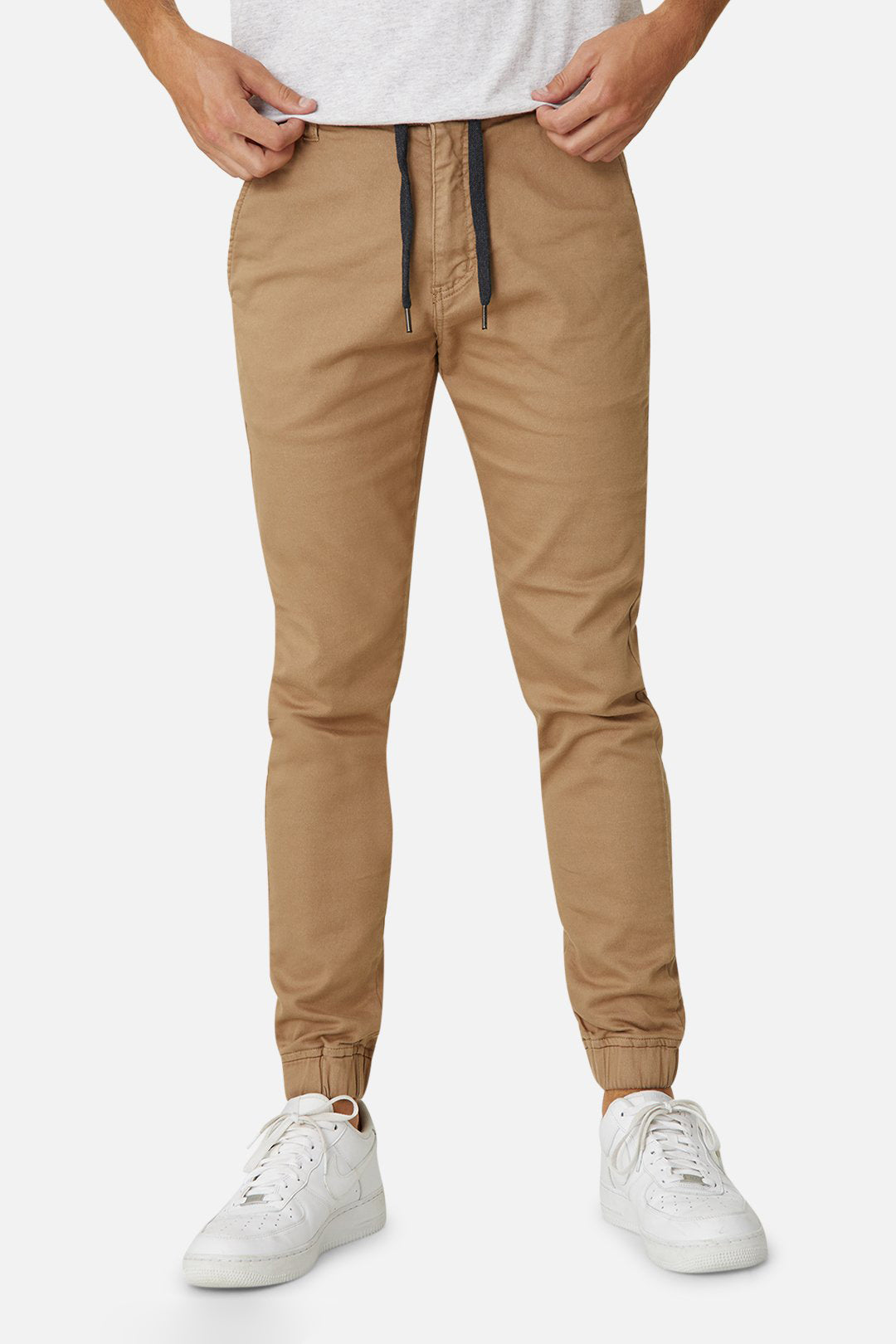 A person wearing Industrie The Drifter Chino Pant in beige with a slim fit and elastic waist, paired with a light gray shirt that's partially visible. They complete the look with white sneakers, all set against a light gray background.