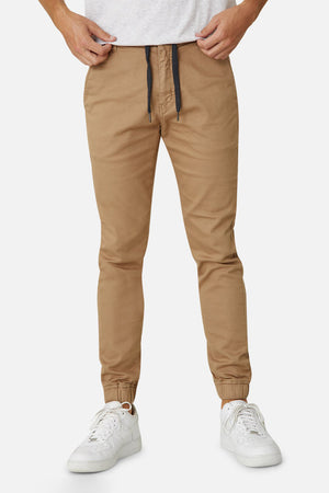 A person wearing Industrie The Drifter Chino Pant in beige with a slim fit and elastic waist, paired with a light gray shirt that's partially visible. They complete the look with white sneakers, all set against a light gray background.