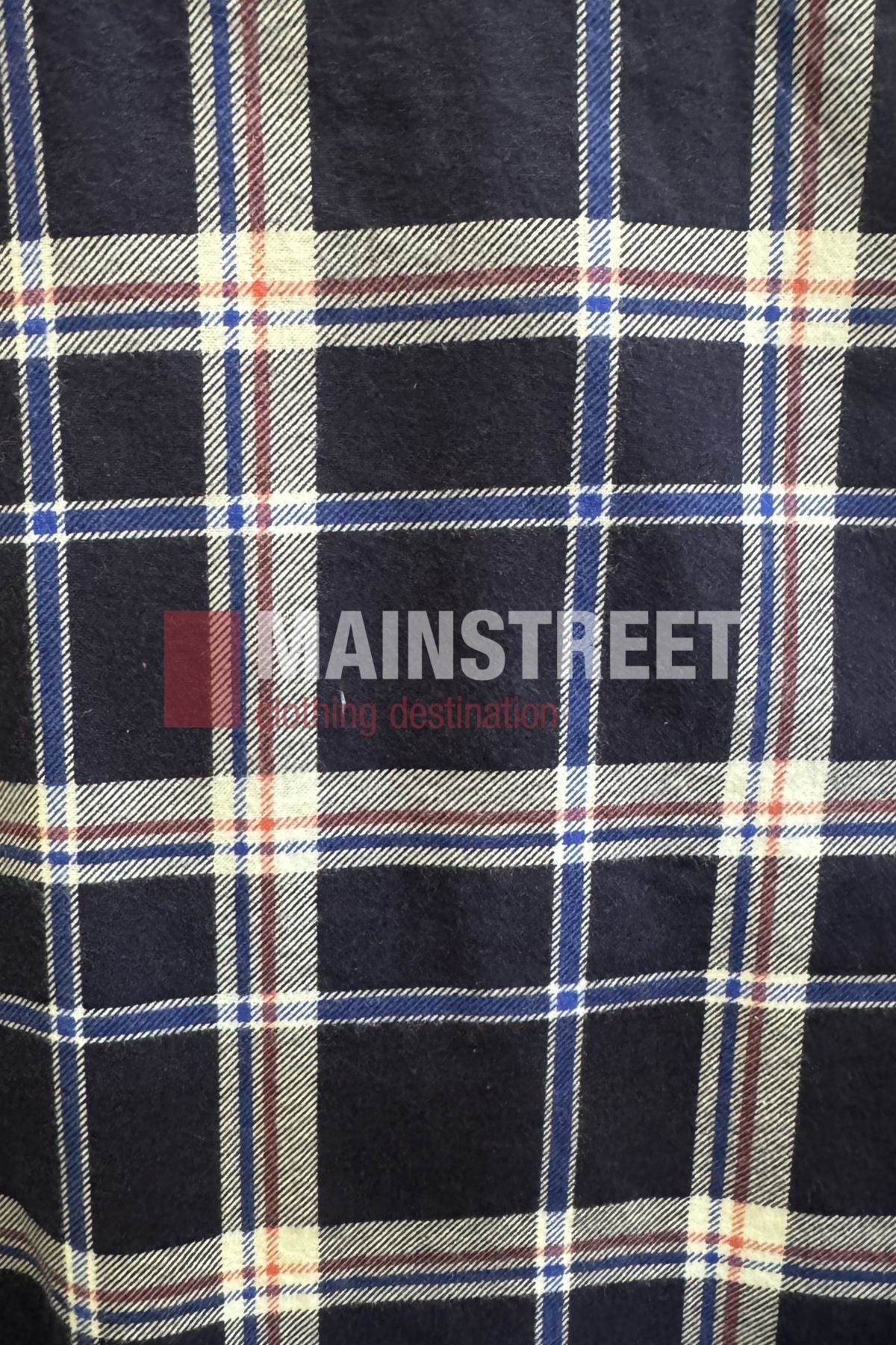 A close-up of navy blue plaid fabric with white, red, and beige lines from the Milton Open Front Flannelette Shirt. "MAINSTREET" and "shopping destination" are partially visible on the extra long tail.