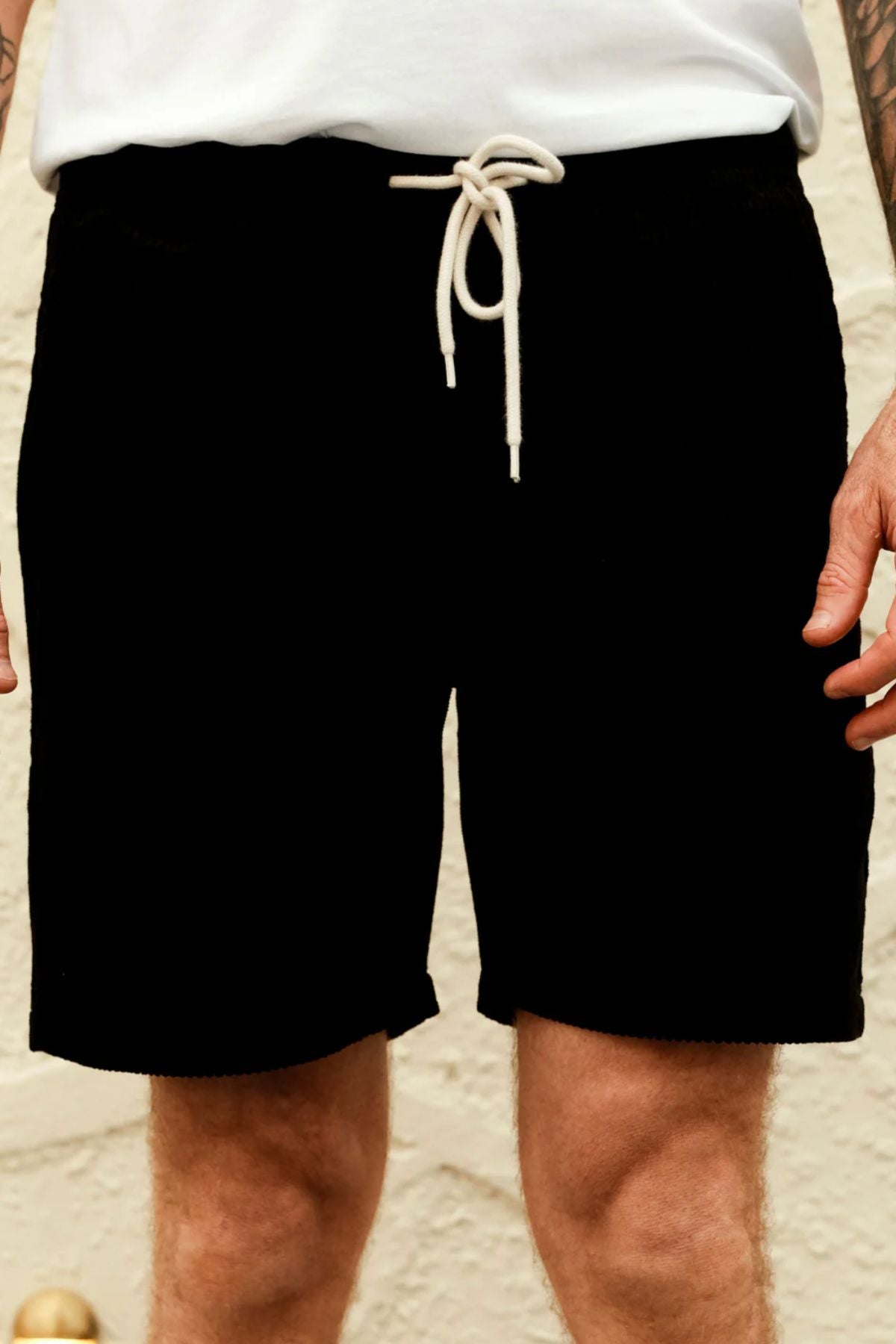 A person wearing Mr Simple Burbank Cord Shorts with a white drawstring and a white T-shirt stands against a light-colored textured wall, with only their lower torso and upper legs visible.