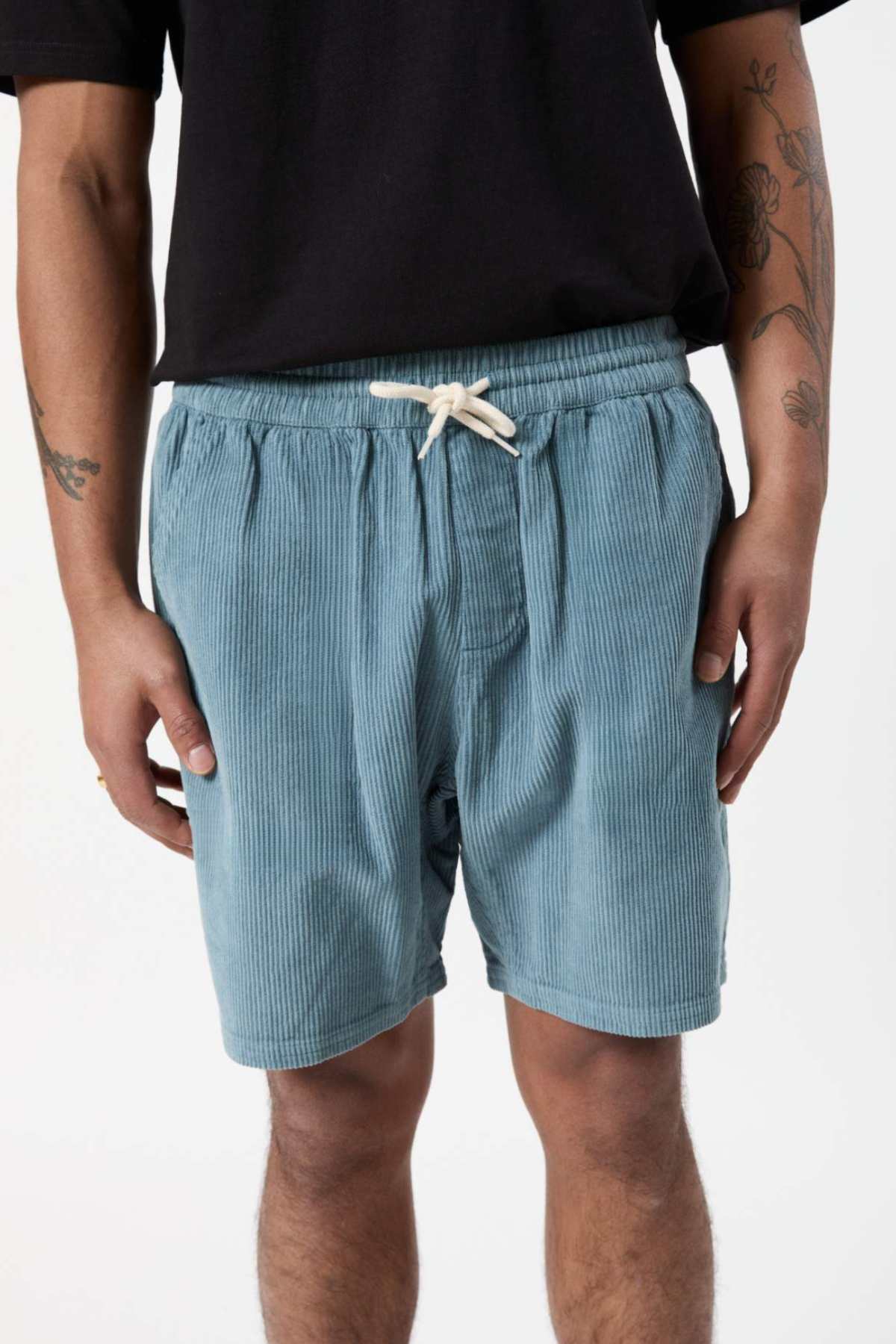 A person wearing light blue Mr Simple Burbank Cord Shorts with a white drawstring stands with their arms by their sides. Sporting a black shirt and visible tattoos on their left forearm, they epitomize the look for a casual outing. The background is plain white.
